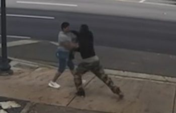 Dos personas se agarran en una acera. Una usa camiseta gris y pantalón azul, la otra lleva ropa oscura y pantalón camuflado. Hay una calle detrás