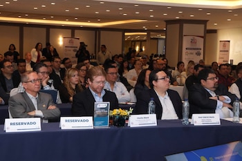Decenas de personas sentadas en un salón de conferencias con mesas cubiertas de tela azul. Hay botellas de agua, carteles con nombres y banners