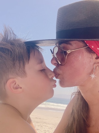 Primer plano de Galilea Montijo con sombrero y gafas de sol besando a su hijo Mateo en la boca, con el mar y la playa de fondo bajo un cielo azul claro