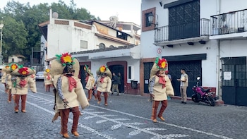 Grupo de personas vestidas con trajes folclóricos, sombreros de paja grandes con flores rojas y amarillas, y máscaras de animales, desfilando por una calle empedrada entre edificios blancos