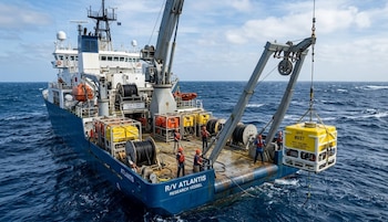 Vista aérea de un gran buque de investigación azul llamado R/V Atlantis con una grúa desplegando un equipo científico amarillo en el océano azul oscuro y ondulado