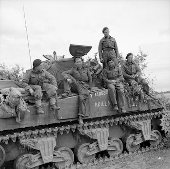 Cinco soldados con uniformes militares y boinas se encuentran sentados y de pie sobre un tanque de guerra verde oliva. El tanque tiene escrito "T 146929 AKILLA" en su lateral y está parcialmente cubierto con vegetación para camuflaje. El cielo está nublado
