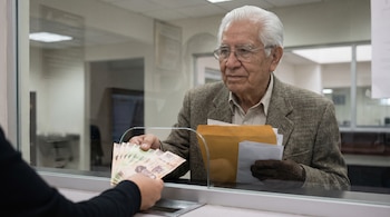 Hombre mayor de cabello blanco y lentes recibe billetes de un cajero a través de una ventanilla de cristal. Sostiene un sobre y documentos.