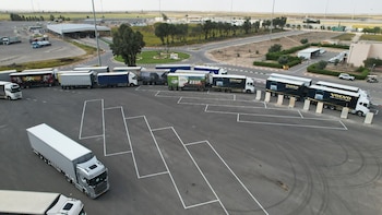 Camiones articulados sobre el asfalto de un estacionamiento con marcas viales y edificios de control al fondo