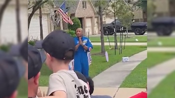 Un hombre con traje azul de astronauta de pie en un césped, hablando con niños cuyas espaldas se ven en primer plano. Detrás, una casa con bandera de EE. UU. y un camión negro