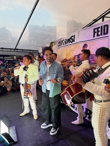 Luis Fonsi en un escenario, sonriente y con micrófono, junto a músicos de mariachi vestidos de blanco tocando instrumentos como violín y trompeta