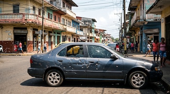 Un automóvil gris con orificios de bala en el costado y cristales rotos permanece en una calle frente a edificios de varios pisos.
