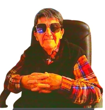 Hombre mayor con cabello gris, gafas de sol oscuras, camisa a cuadros y chaleco oscuro, sentado en una silla con las manos entrelazadas
