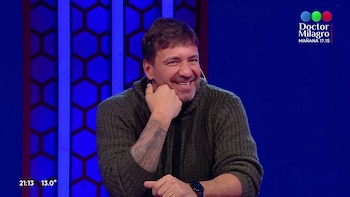 Un hombre sonriente con suéter verde y tatuajes en el brazo apoya la barbilla sobre su mano en un estudio de televisión con fondo azul