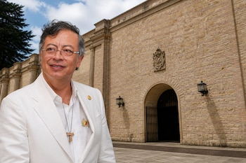Gustavo Petro con traje blanco, anteojos y crucifijo de madera frente a un edificio de piedra con puerta en arco.