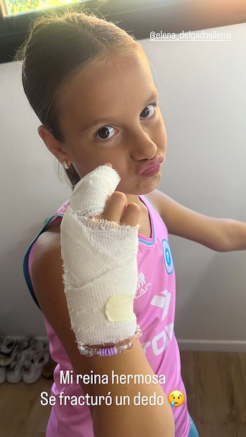 Primer plano de una niña con camiseta rosa, mostrando su mano derecha con un dedo vendado. Tiene los labios fruncidos y mira directamente a la cámara