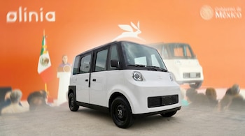 Un vehículo eléctrico compacto blanco con techo y ruedas negras está estacionado sobre suelo de concreto, con un fondo naranja difuminado con logos y figuras humanas.