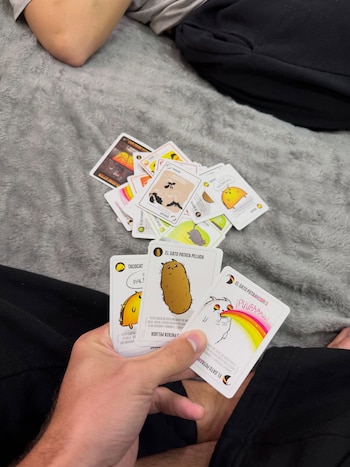 Mano de una persona sosteniendo tres cartas del juego Exploding Kittens, con más cartas esparcidas sobre una manta de color gris