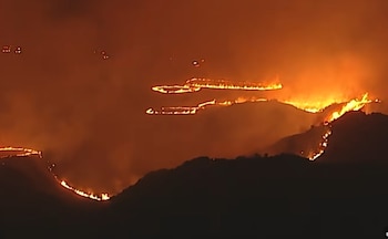 Vista aérea nocturna de un incendio forestal masivo, con brillantes líneas de fuego avanzando sobre varias crestas montañosas oscuras bajo un cielo naranja rojizo