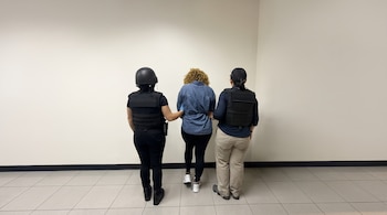 Tres personas de espalda frente a una pared blanca: dos agentes con chaleco táctico sujetan a una mujer con camisa de mezclilla y esposas.