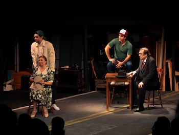 Cuatro personas en un escenario de teatro iluminado con sillas, una mesa pequeña de madera y utilería