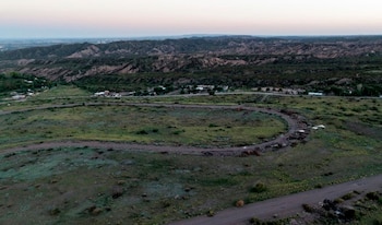 Vista aérea de un paisaje semidesértico con una pista de tierra curva, un camino recto y edificios dispersos; montañas y cielo claro en el fondo