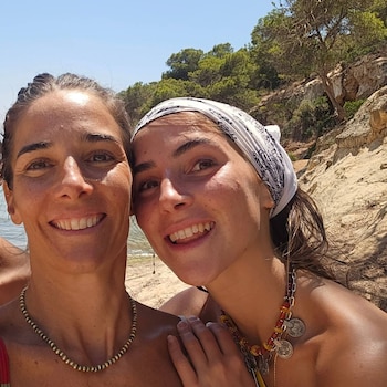 Dos mujeres sonríen a cámara, una con pañuelo blanco estampado y collar de monedas. El fondo muestra arena, mar, rocas y árboles