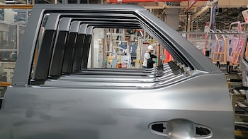 Vista interior de una fábrica de automóviles con múltiples puertas de vehículos sin pintar en primer plano, y un operario con casco y guantes trabajando al fondo