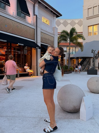 Camila Homs sonríe mientras sostiene en brazos a una niña pequeña frente a tiendas en un paseo peatonal al aire libre