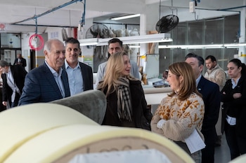 Varias personas en una fábrica con rollos de material de espuma en primer plano, luces fluorescentes en el techo y un ventilador en el fondo