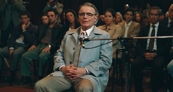 Un hombre con gafas, gabardina gris y corbata estampada está sentado con las manos cruzadas frente a un micrófono, rodeado de otras personas