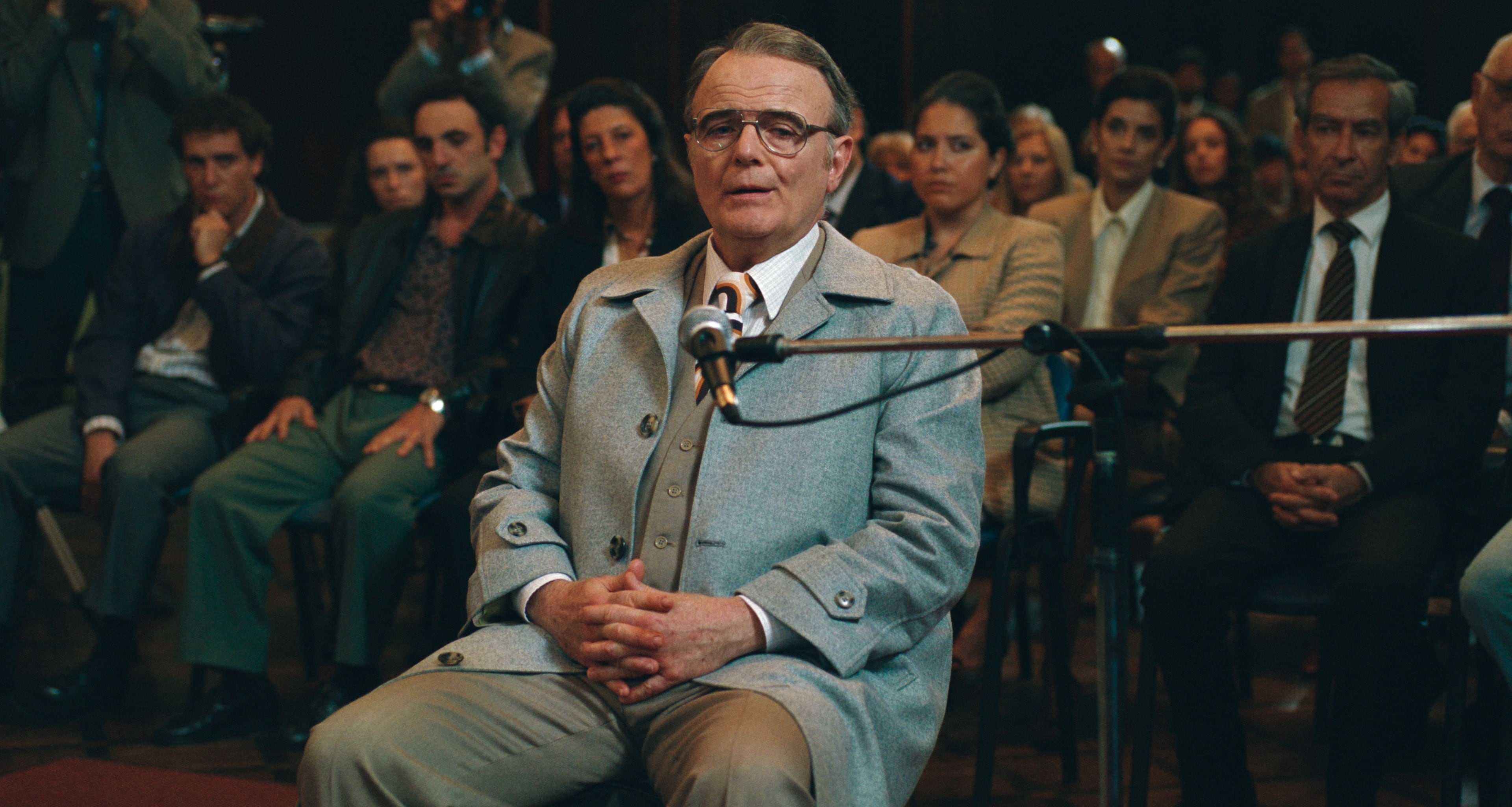 El actor Luis Machín, caracterizado como Ricardo Barreda, se sienta frente a un micrófono en una sala de audiencia, con varios asistentes detrás. (Prime Video)