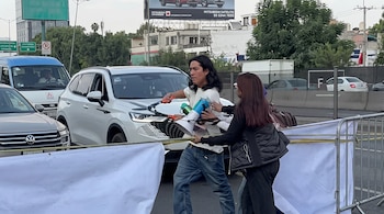 Un hombre con megáfono y una mujer caminan junto a un automóvil blanco sostenidos detrás de una lona blanca y una valla metálica.