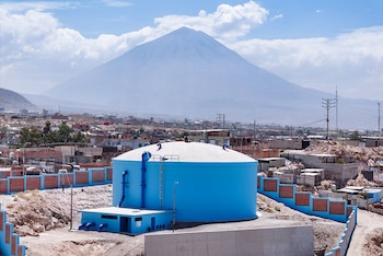 Un gran tanque circular de agua pintado de azul junto a viviendas y un volcán cónico de gran tamaño al fondo bajo un cielo con nubes