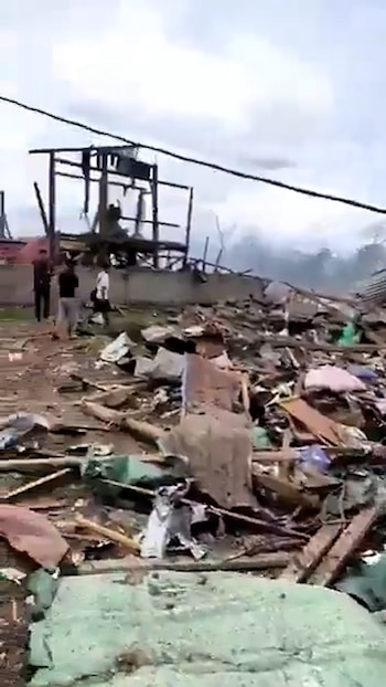 Un paisaje devastado por una explosión, con montones de escombros y restos de un edificio destruido en el primer plano y fondo. Varias personas observan la destrucción