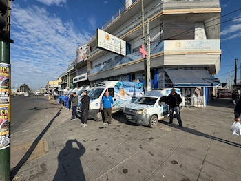 Cinco vehículos de servicio de Argentina Salud (vans y un coche subcompacto) estacionados en una acera urbana. Personal junto a ellos, con edificios y calle de fondo bajo cielo azul