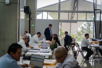 Varias personas están sentadas frente a mesas con ordenadores y carpetas en un espacio con amplios ventanales