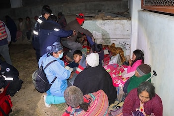Personas sentadas en el suelo cubiertas con mantas, algunas con uniformes de emergencia, un paramédico con barbijo. Entorno exterior oscuro con una pared de fondo