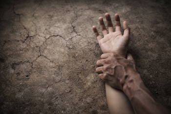 Una mano fuerte sujeta la muñeca de otra mano más clara, ambas sobre una superficie de tierra seca y agrietada.