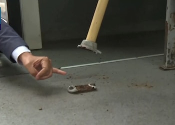 Una mano señala una pieza de metal oxidada sobre un suelo de concreto gris. También se ve parte de un poste con pintura descascarada y un objeto con mango de madera claro