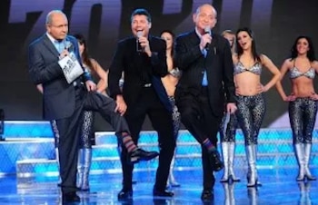 Tres hombres en traje con micrófonos están en un escenario brillante junto a bailarinas, mientras dos de ellos muestran sus calcetines