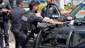 Varios agentes de policía uniformados rodean un vehículo policial Ford negro con rejilla delantera. Una bolsa negra y una gorra están sobre el capó