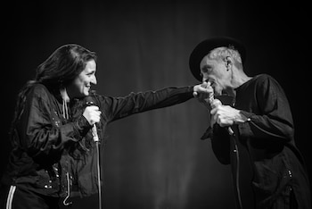 Fotografía en blanco y negro de una mujer y un hombre con sombrero que sostienen micrófonos y se saludan sobre un escenario