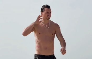 Hombre de cabello oscuro peinado hacia atrás, sin camiseta, con collar plateado, tatuajes en el torso y brazo. Sonríe levemente y se toca la cabeza