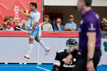 Un jugador con uniforme de hockey blanco y celeste salta con un palo en la mano frente al público y a un arquero en el suelo