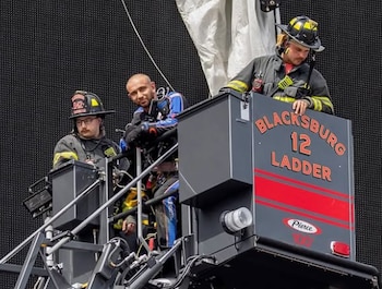 Tres hombres en la plataforma elevada de una escalera de camión de bomberos de Blacksburg. Dos llevan uniformes de bombero; el central viste un mono deportivo azul y negro