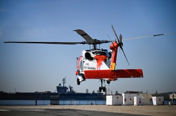 Un helicóptero blanco y naranja vuela a baja altura sobre una pista cerca de un muelle con un buque militar y el cielo despejado
