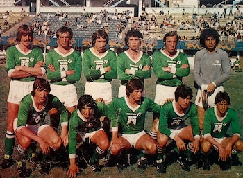 Once jugadores de fútbol con uniformes verdes, blancos y grises posan en fila, algunos de pie y otros de rodillas, en un campo de césped frente a gradas con público