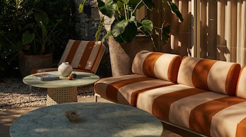 Un sofá y un sillón a rayas naranjas y beige en un patio con dos mesas redondas de mármol, plantas en macetas grandes y luz solar.