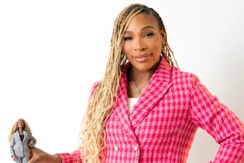 Serena Williams, con un blazer fucsia de pata de gallo, sostiene una muñeca Barbie que usa un traje gris de dos piezas y cabello trenzado rubio