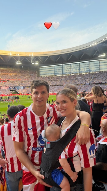 Gianluca Simeone y Eva Barigela sonríen a cámara con su bebé Faustino en un portabebés, vistiendo camisetas rojiblancas del Atlético de Madrid en un estadio