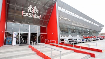 Entrada de un edificio moderno con letreros de EsSalud e INCOR, rampas de acceso rojas, escaleras y vehículos en el estacionamiento.