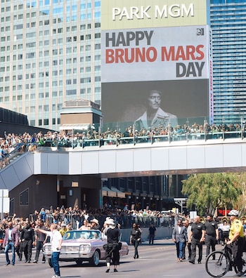 Calle de Las Vegas con edificios, multitud en la acera y un puente peatonal, un cartel gigante de 'Día de Bruno Mars' y un desfile con un coche rosa
