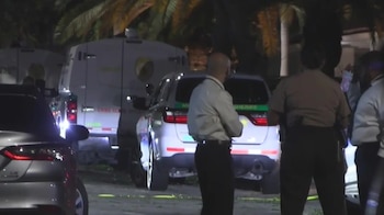 Vehículos de la policía y forenses, incluyendo una furgoneta de "Crime Scene" y un coche de "Sheriff", en una calle oscura con oficiales y palmeras al fondo