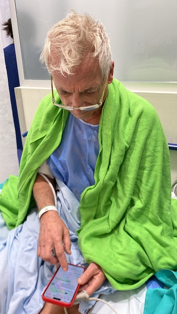 Hombre rubio canoso, con gafas en la frente, cubierto con una manta verde, sentado en una camilla hospitalaria, usando su teléfono. Lleva una bata azul y una pulsera